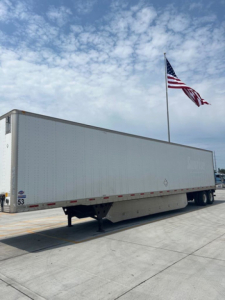 Utility van trailer with a white box, landing gear visible, and an American flag in the background. Side of trailer shows no readable branding.