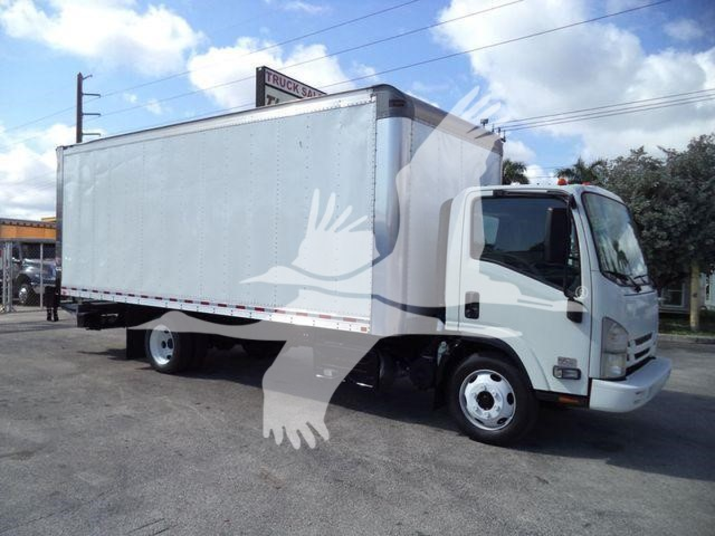 Isuzu box truck with a large white cargo box mounted on a cab chassis; text on sign in background blurred; no legible model details visible on the truck.