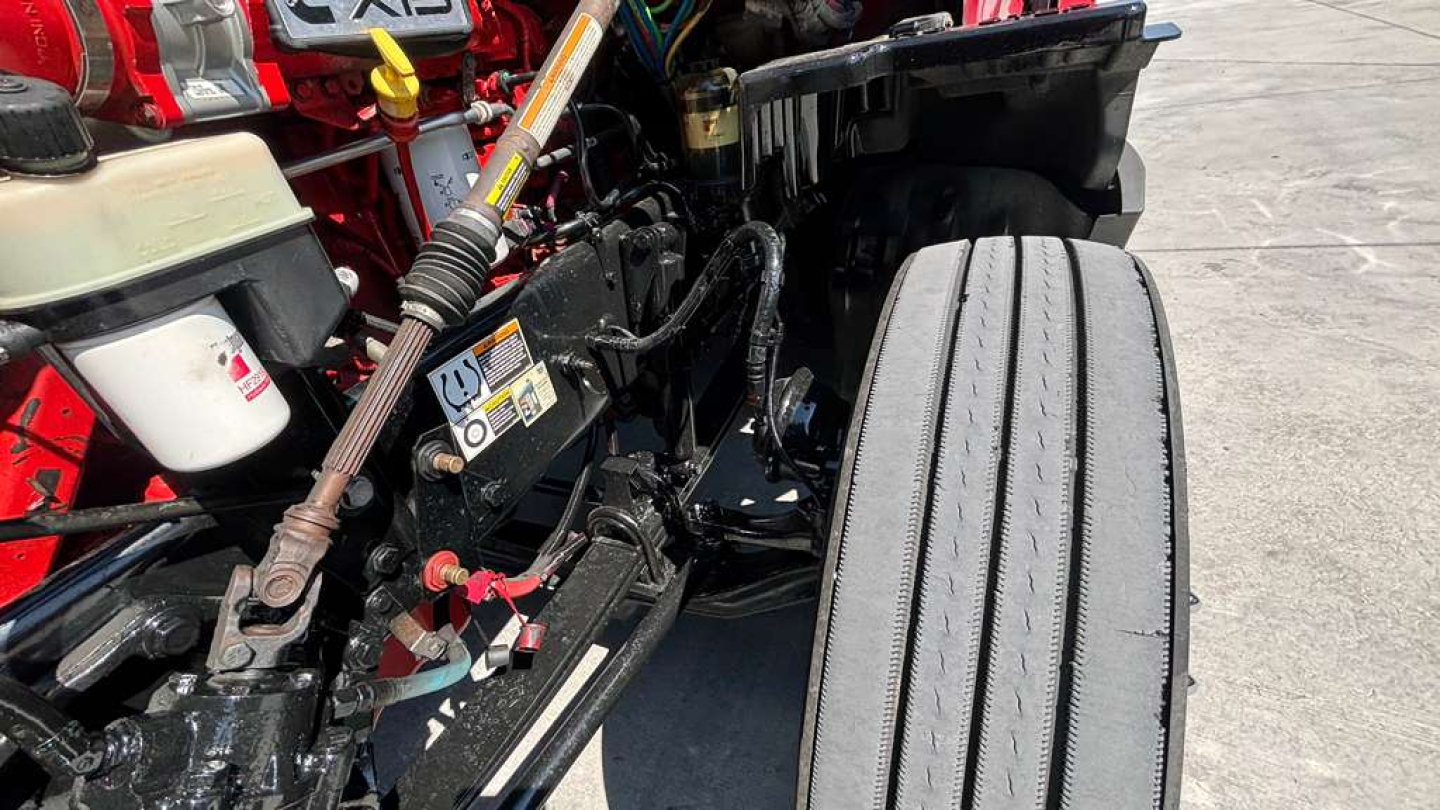Kennworth T680 engine bay area with visible hydraulic lines, warning label, and a front tire; various components including a white reservoir and red chassis nearby.