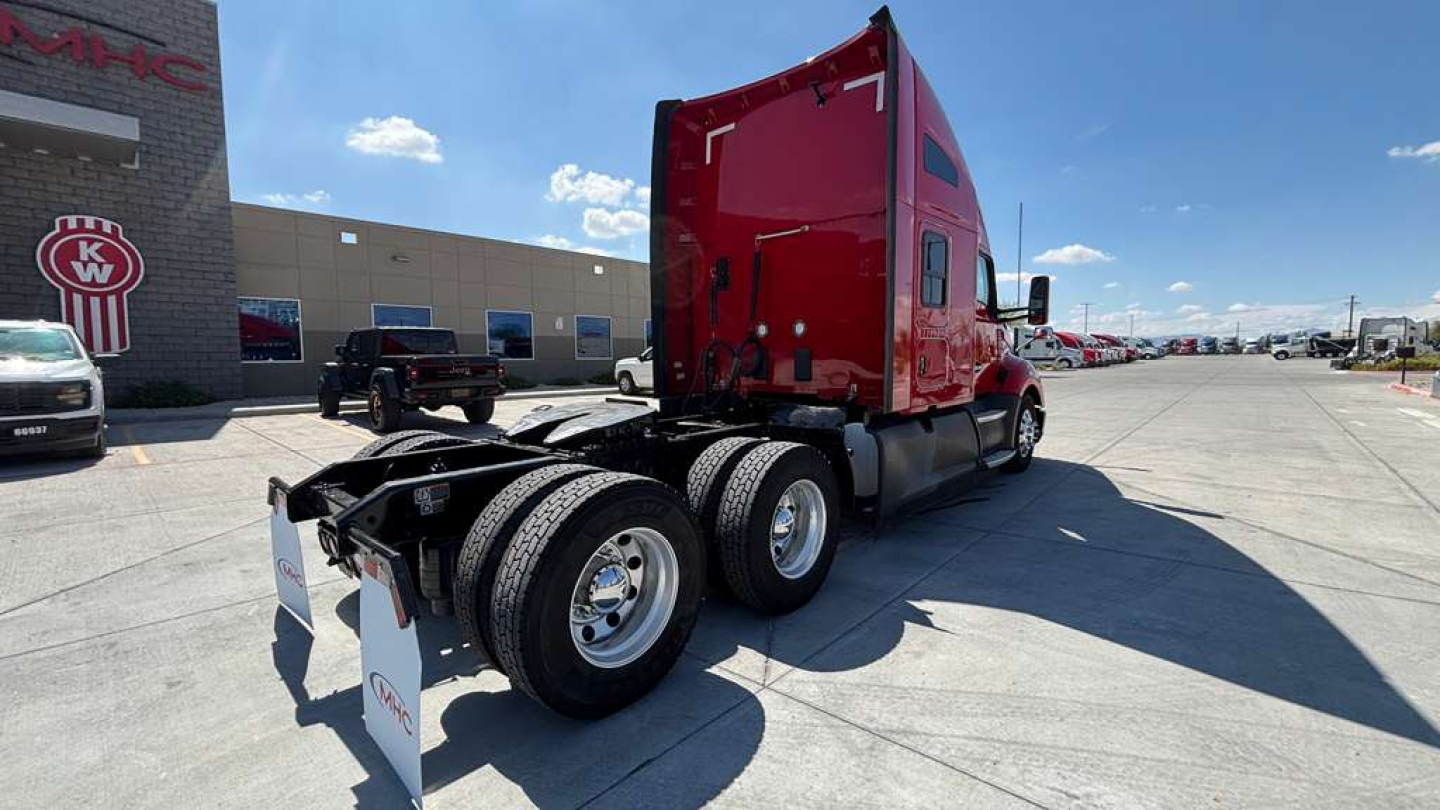 Kenworth T680 semi-truck in a dealer lot, red sleeper cab, rear view showing triple axles and dual rear tires, registration and stock number on sign in foreground