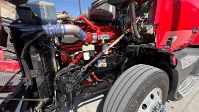 Kenworth T680 engine bay with exposed front suspension and turbo piping, visible VIN label area, various hoses and red cab, front left wheel and fender.