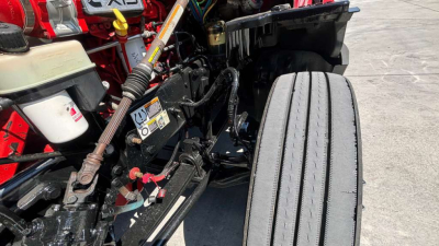 Kennworth T680 engine bay area with visible hydraulic lines, warning label, and a front tire; various components including a white reservoir and red chassis nearby.