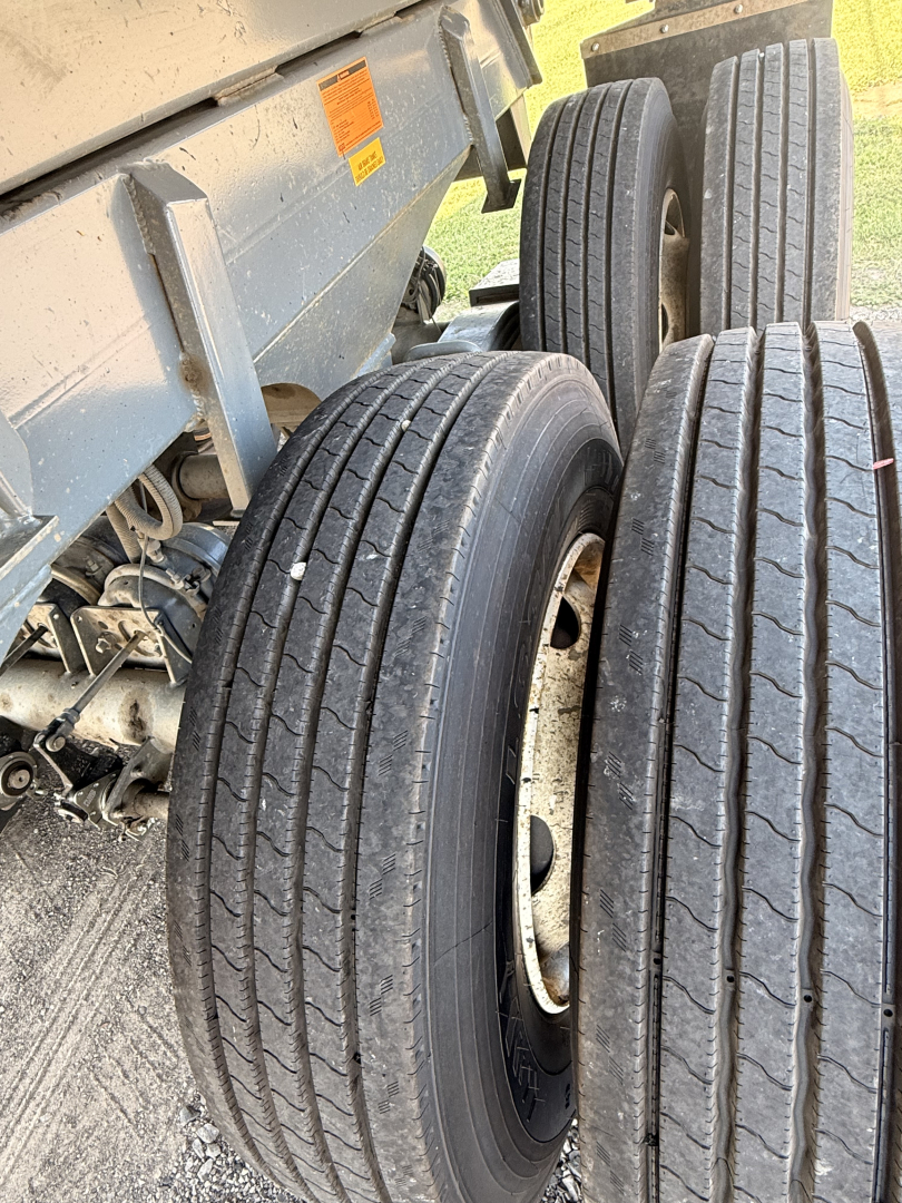 Trailer wheel assembly with dual axles and multiple tires visible; metallic chassis and orange warning sticker on the side of the dump trailer; treaded tires and rims exposed.