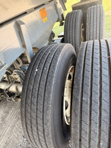 Trailer wheel assembly with dual axles and multiple tires visible; metallic chassis and orange warning sticker on the side of the dump trailer; treaded tires and rims exposed.
