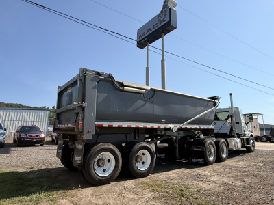 Segment of a dump trailer with a gray Ranco end dump bed. Dual rear axles with six wheels visible, and a Long 3-axle tractor in the background. The side of the bed shows no legible labels clearly visible in this view.
