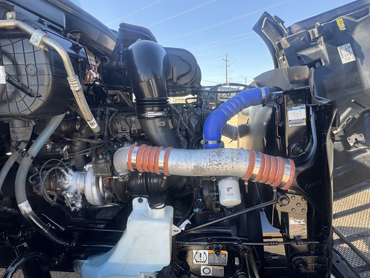 Peterbilt 579 engine bay with turbocharger, intercooler piping, coolant reservoir, and blue silicone hose visible. Engine components and surrounding framing show under-hood layout.