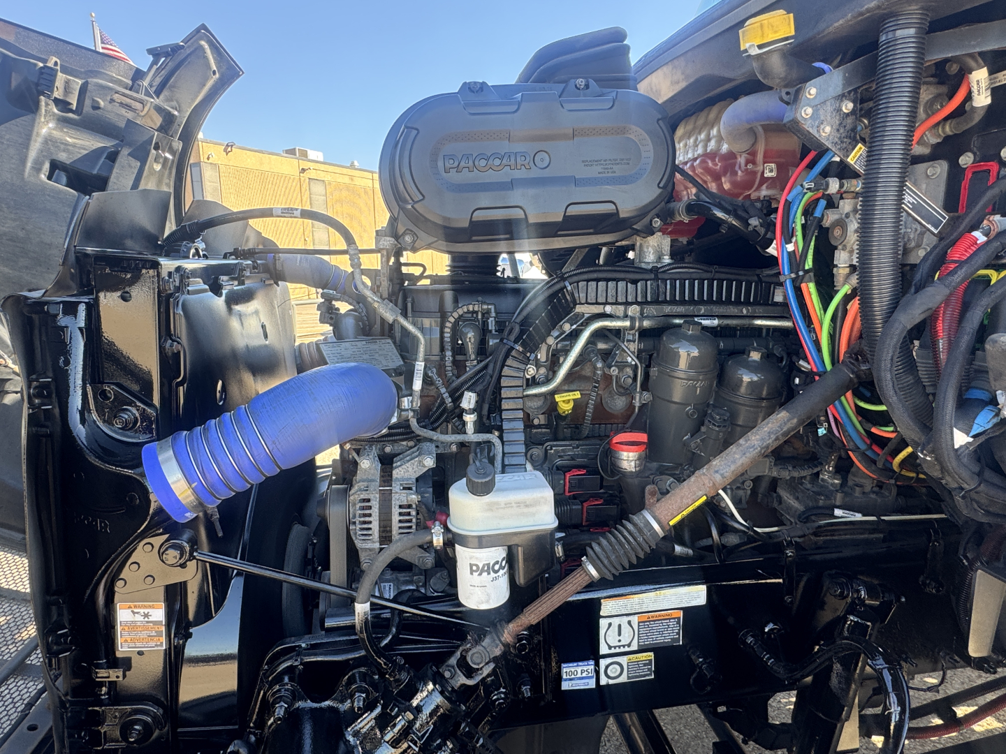 Truck engine bay of a 2022 Peterbilt 579 showing air intake, hoses, wiring, and fluid reservoirs including an angled white coolant/def reservoir and visible labels.