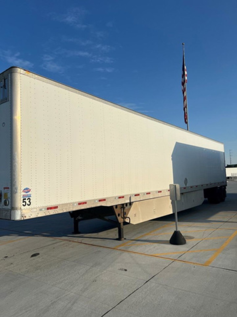 Rear-left view of a white utility van trailer with a support leg, raised landing gear, and a circular emblem near the front; blue sky overhead.