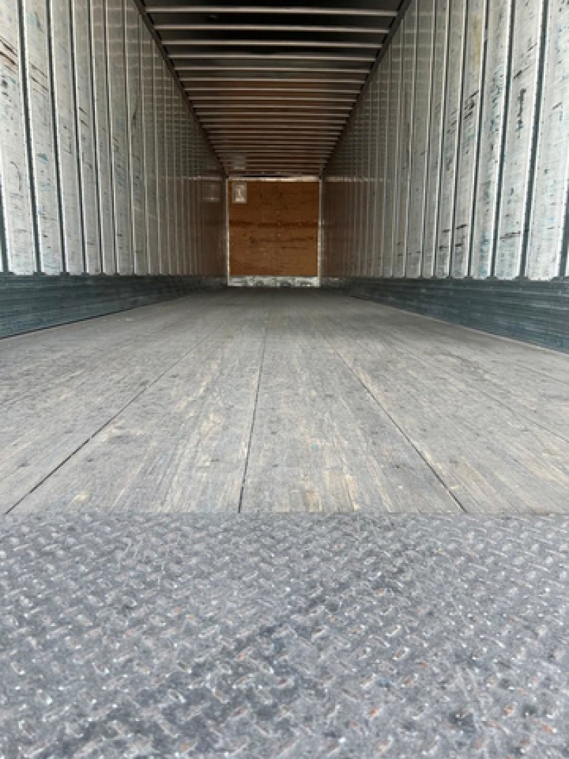 Trailer interior showing empty van trailer with wooden bulkhead at far end and metal ribbed walls; textured floor near camera.