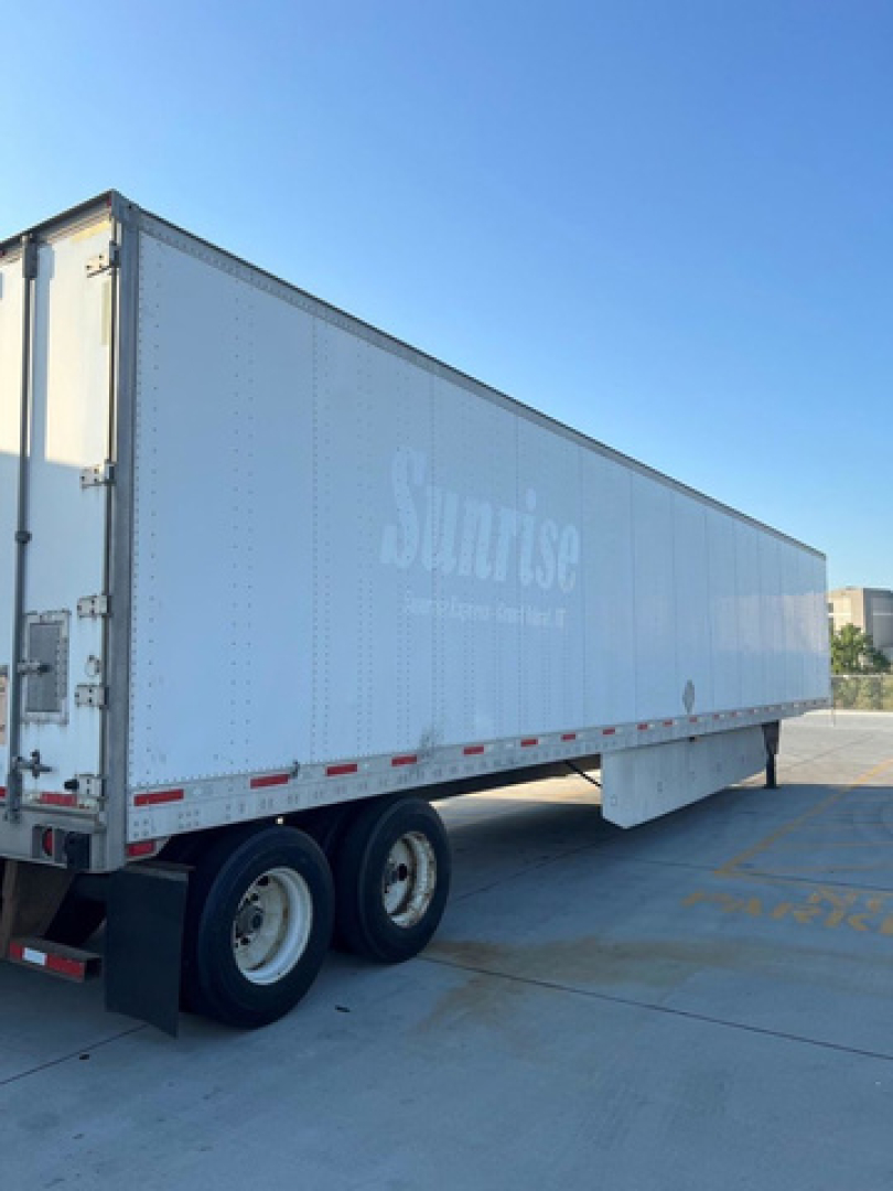 Trailer side of a white SunRise trailer with faint lettering; dual rear wheels; boxed trailer in a parking lot.