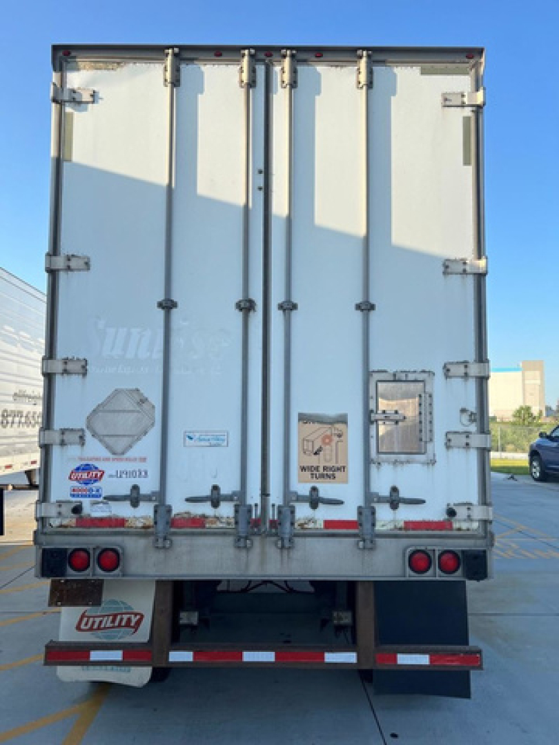 Rear view of a white utility van trailer with double doors, various reflective tape, and stickers including warning placards and a Utility brand mudflap.