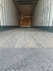 Trailer interior showing empty van trailer with wooden bulkhead at far end and metal ribbed walls; textured floor near camera.