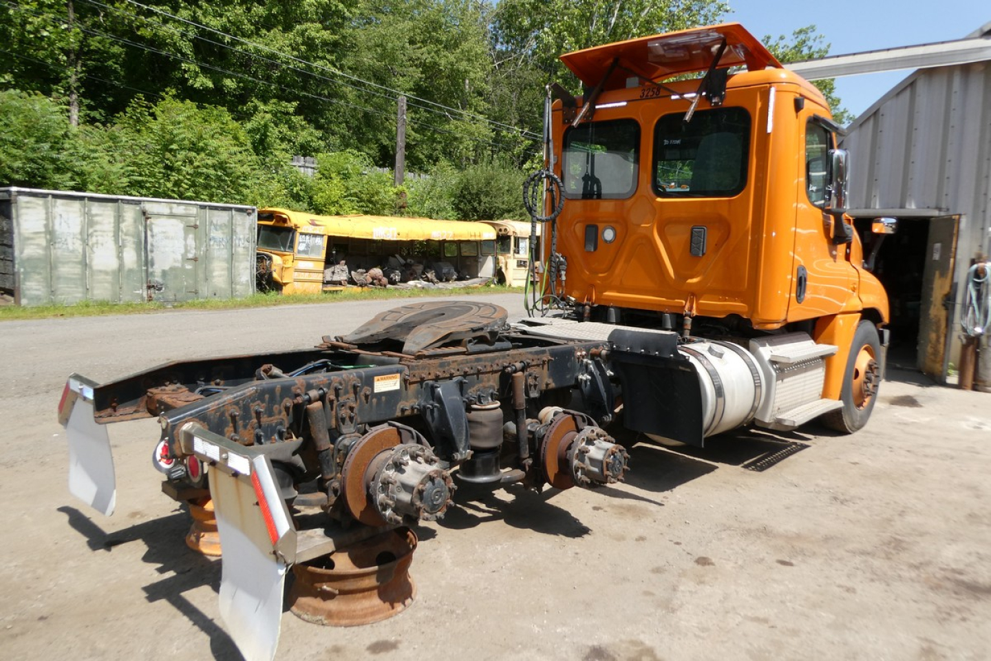 Orange Freightliner Cascadia truck cab with missing trailer chassis and exposed axles. Front of cab visible with windows and mirrors; rusty undercarriage components.