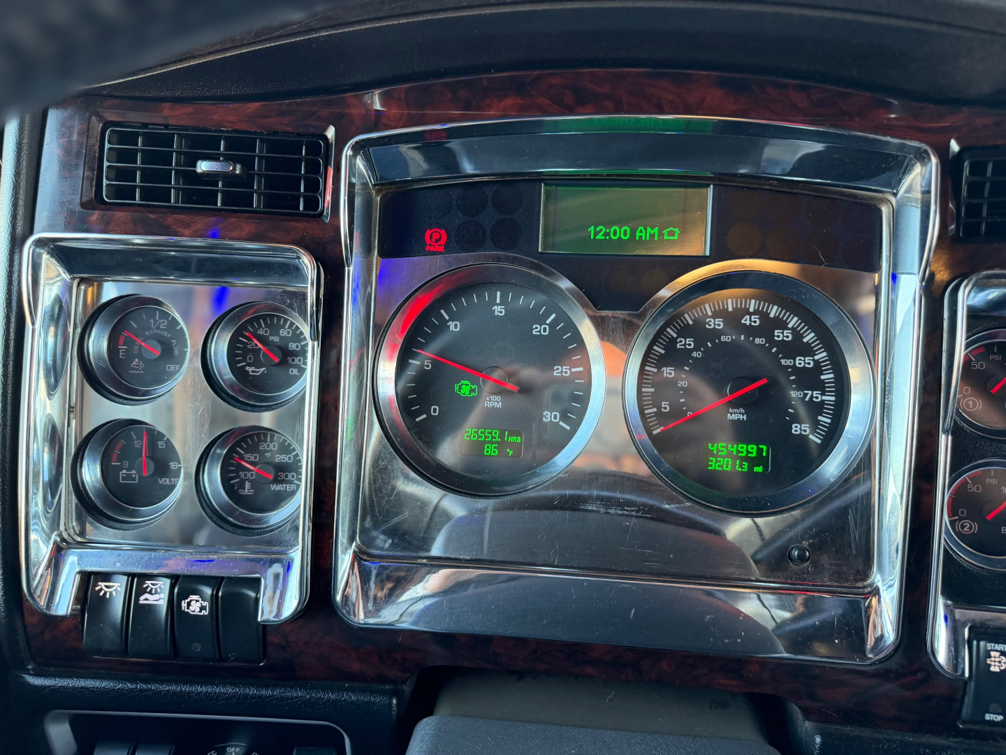 Kenworth W900L dash cluster showing multiple gauges and center digital clock, with woodgrain trim and chrome surroundings.
