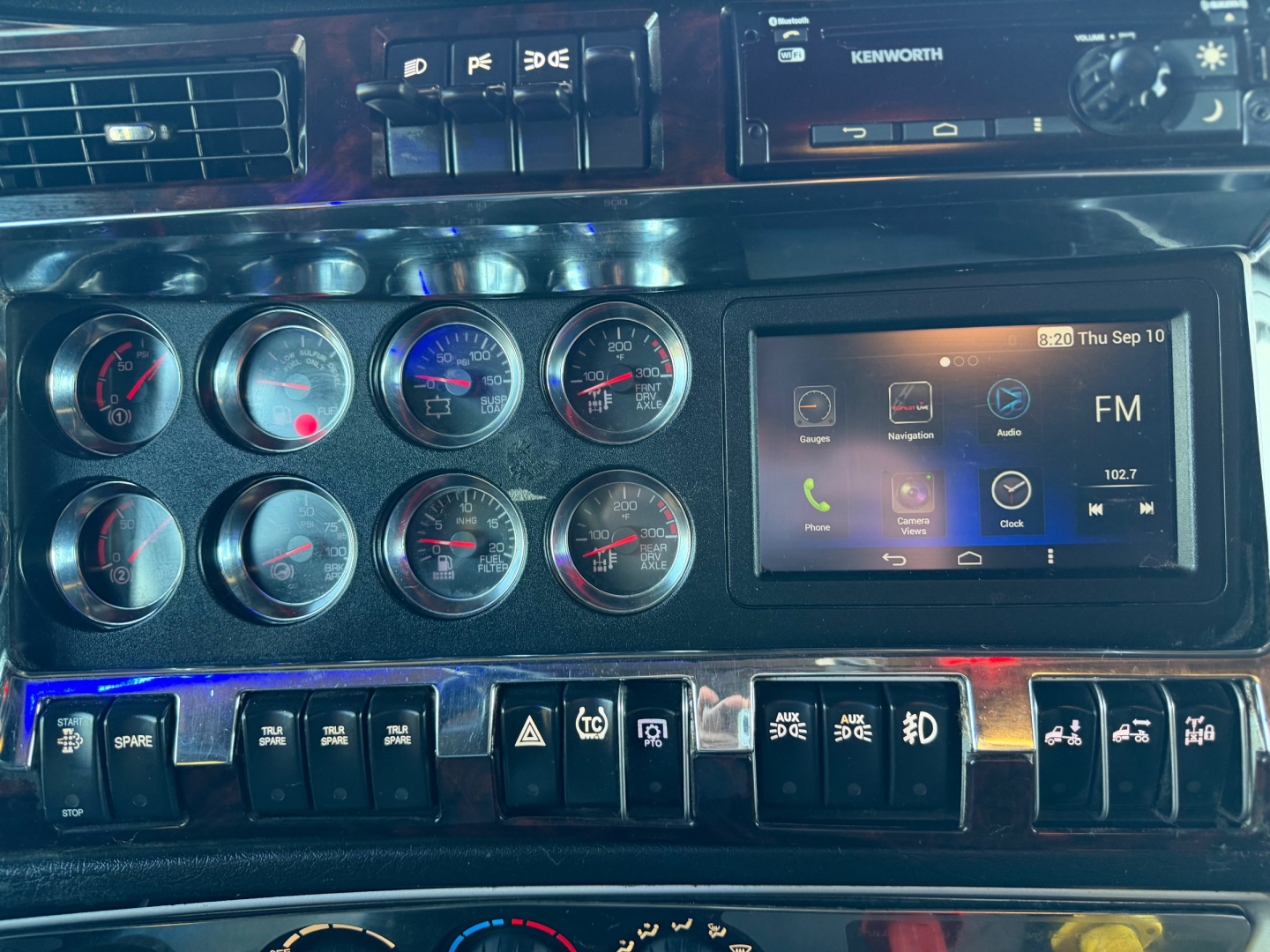 Kenworth W900L dash panel showing multiple round gauges, a Kenworth-branded radio/navigation screen, and various control switches.