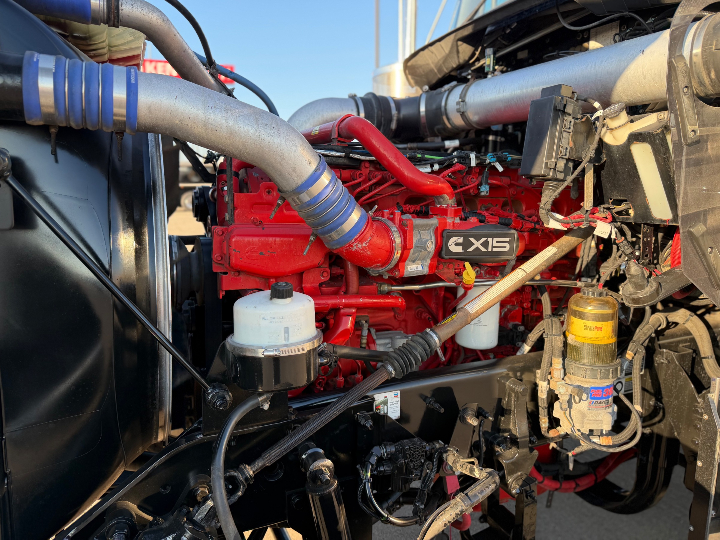 Truck engine bay of a 2021 Kenworth W900L showing a red engine with radiators and hoses; visible labels include 'C15' on the engine, white coolant reservoir, yellow oil filter, and various hoses.
