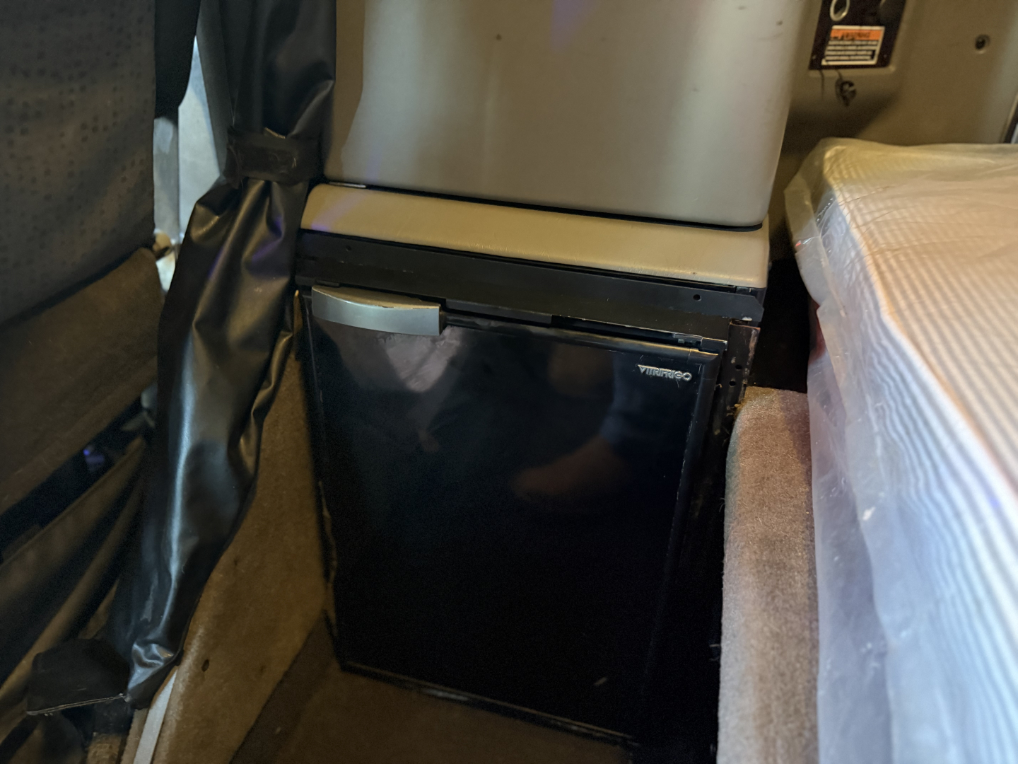 Kenworth W900L interior showing a compact black fridge/freezer unit under the dash between the seat and bed area.