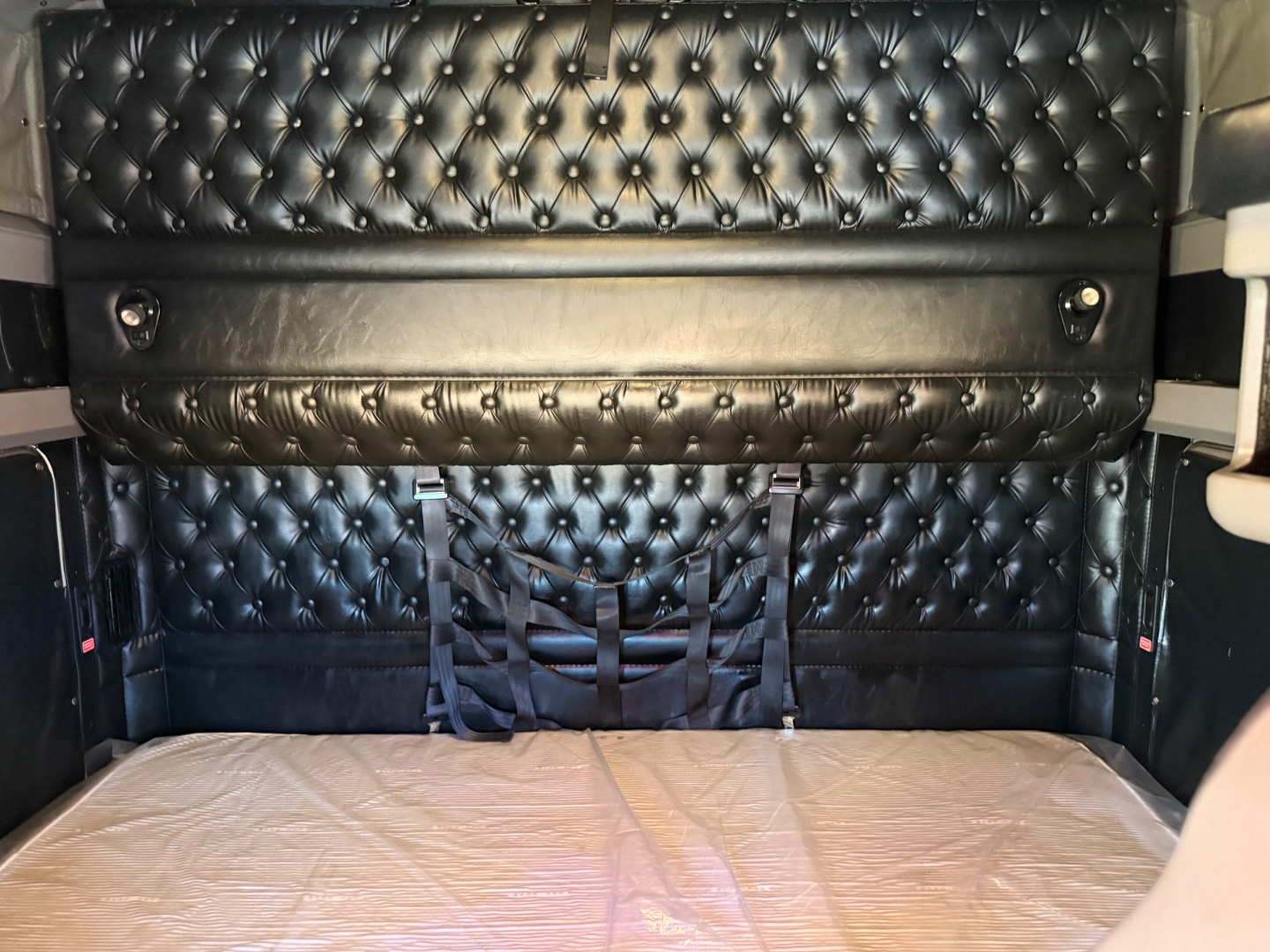 Truck sleeper area with quilted black leather upholstery and a sunken bed platform. Visible straps hang from a small net pocket and a padded shelf/strap along the wall.