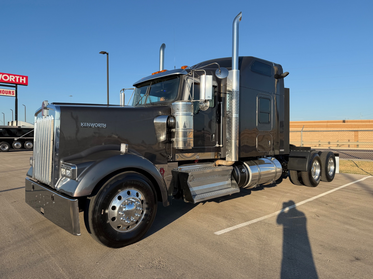 Kenworth W900L semi-truck in a parking lot, glossy black sleeper cab with chrome exhaust stacks and polished wheels, front view angled to the left.