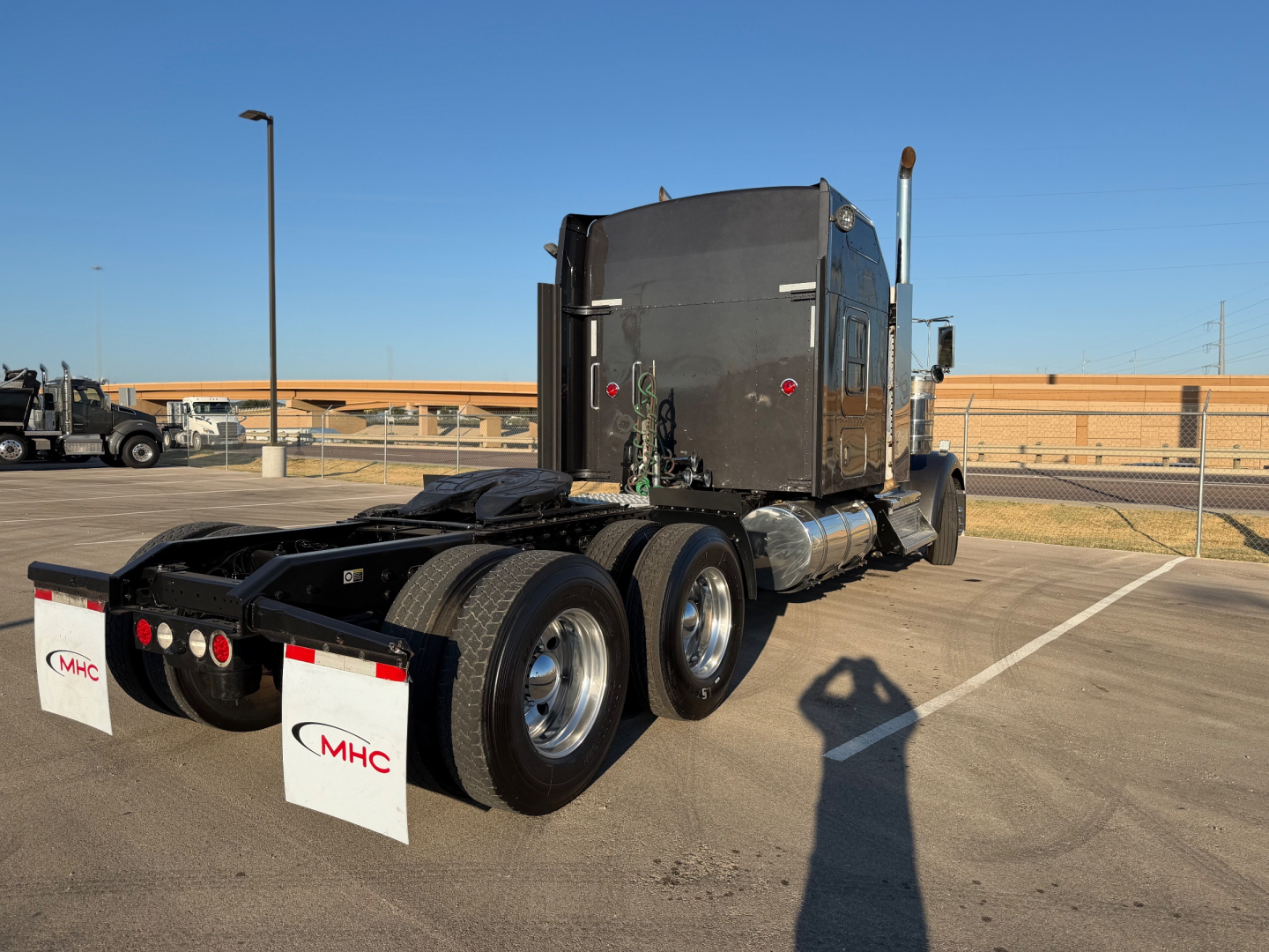 Kenworth W900L truck tractor in a lot, rear three-quarter view, tandem axles, sleeper cab, fuel tanks, mud flaps with MHC logo.