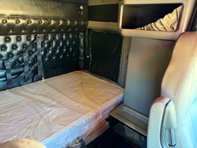 Kenworth W900L sleeper cab interior with quilted black padded wall, single bunk bed with plastic mattress, storage shelves, and beige seat; compact workspace visible.