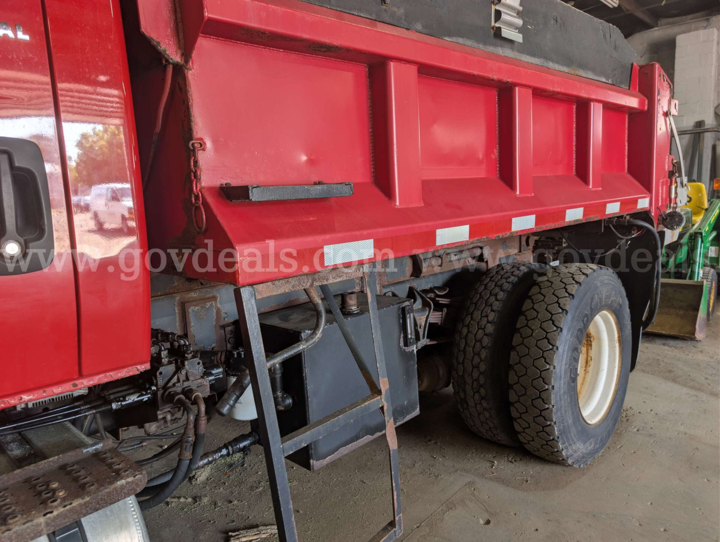 Dump truck side view showing red dump bed and dual rear wheels with white rims, hydraulic lines, and undercarriage components.