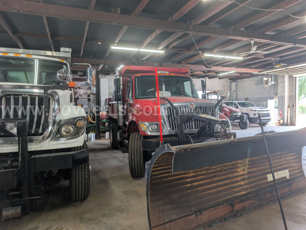 Truck bay with a red International 7400 dump truck in the center, flanked by other trucks and a large snow plow blade in the foreground.