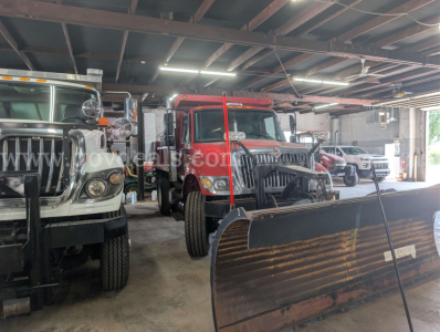 Truck bay with a red International 7400 dump truck in the center, flanked by other trucks and a large snow plow blade in the foreground.