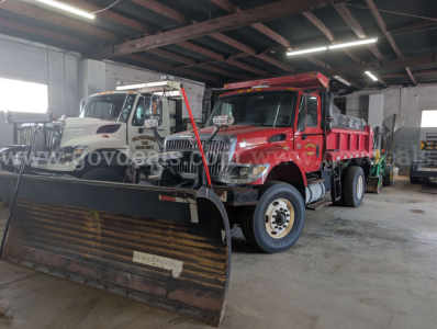 Two heavy-duty dump trucks inside a warehouse; red dump truck in front with snow plow attachment and white dump truck behind.