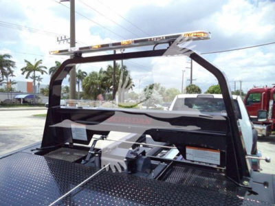 Ram 5500 rollback truck bed with steel diamond plate surface, rear stake rack, and cab protection panel; visible label text includes RAM branding on panel and a rooftop light bar.