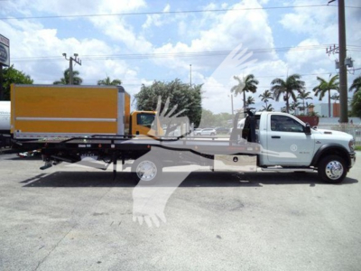 Ram 5500 rollback truck side view with white cab and yellow dump bed, flatbed trailer, palm trees and blue sky in background.