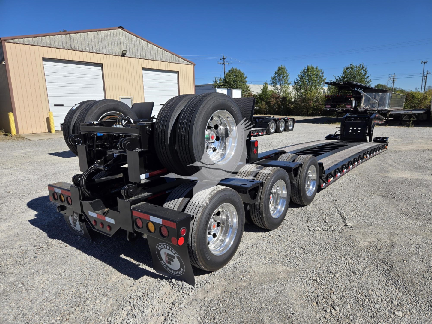 Trailer rear view showing multi-axle Fontaine lowboy with flip axle hydraulic system and spare tires mounted, black chassis, gravel lot, industrial building in background.