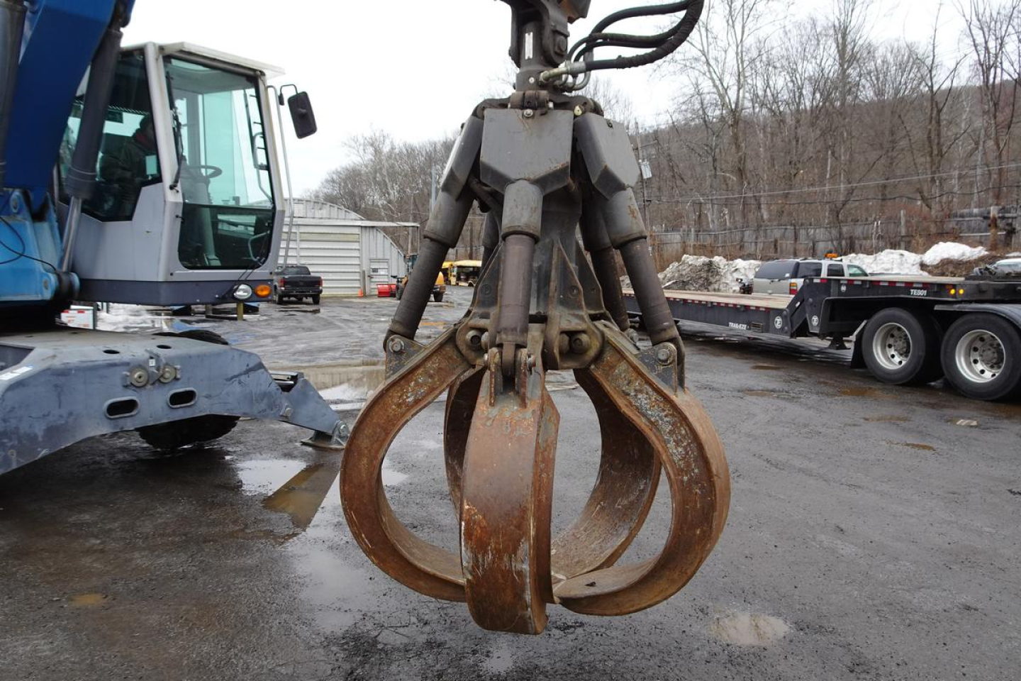 Terex MHL 331 clamshell grapple attachment with rusted steel claws and hydraulic lines in foreground, industrial yard in background.