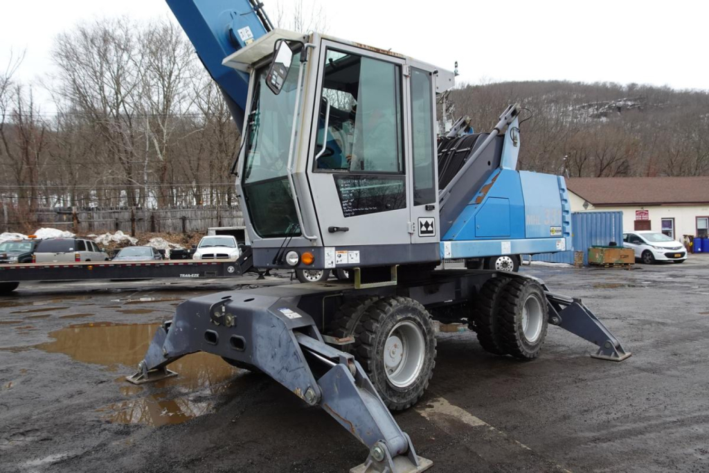 Terex MHL 331 mobile crane in a parking lot, side view showing blue lifting arm, operator cab, and stabilizers deployed on ground.
