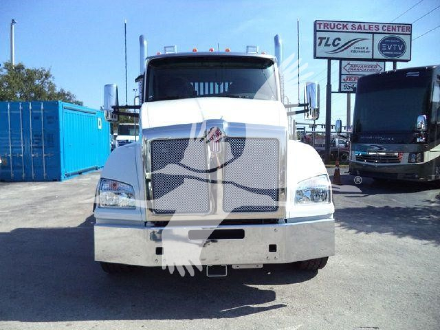Kenworth T880 front view in a dealership lot with chrome grille, headlights, and bumper; blue containers and another truck in background.