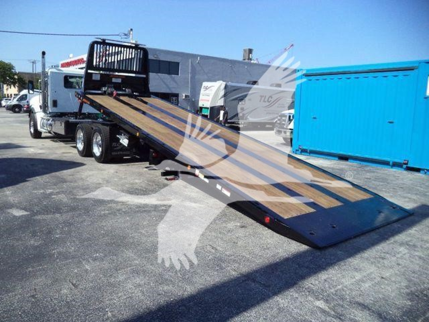 Kenworth T880 rollback truck with a long, lowered steel tilt-bed trailer in a parking lot; blue portable office in background; stock number J68214 visible on listing signage
