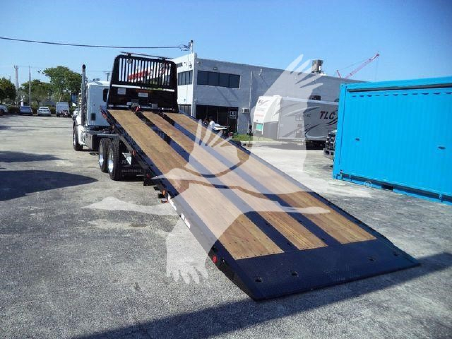 Kenworth T880 rollback truck with a long wooden ramp extending from the rear, parking lot with storage containers and other trucks.