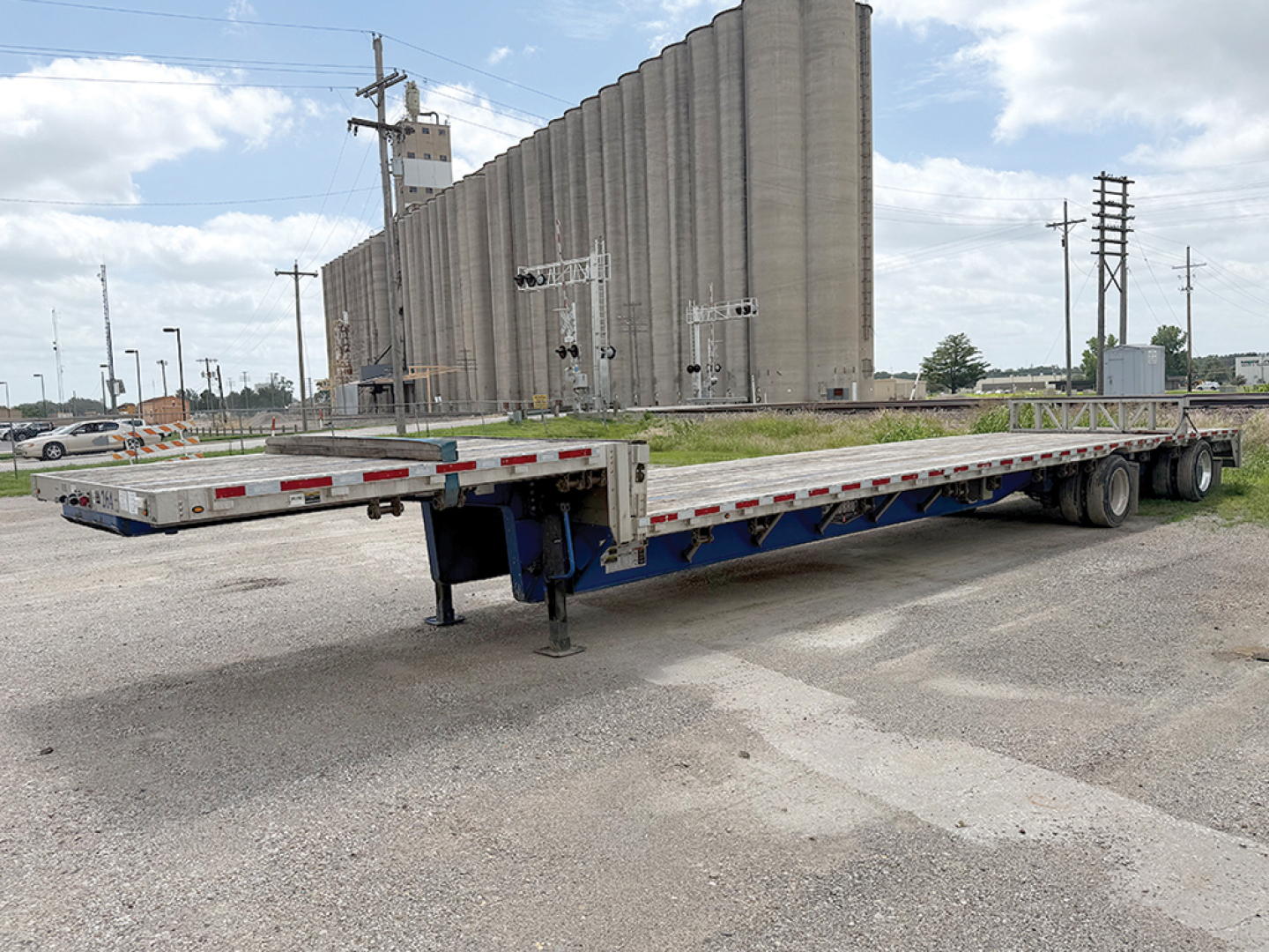 Trailer showing a long flatbed trailer in front of a grain silo with a blue frame and landing gear deployed.