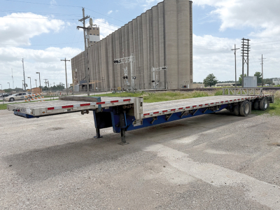 Trailer showing a long flatbed trailer in front of a grain silo with a blue frame and landing gear deployed.