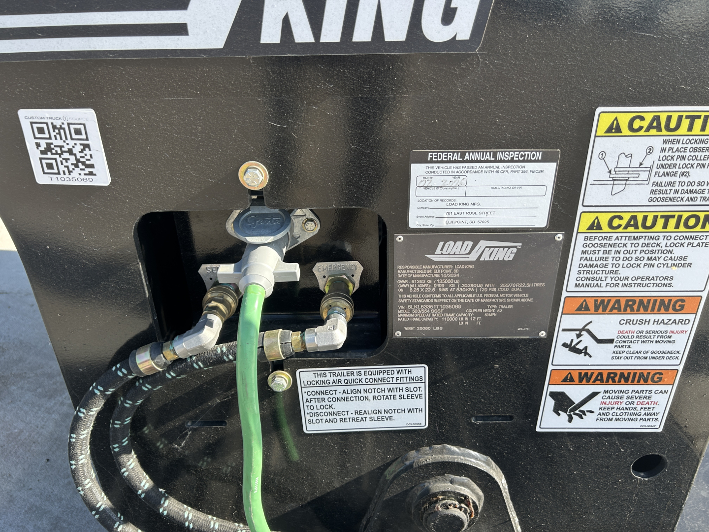 Lowboy trailer back corner with labels and hoses. Visible text includes CAUTION/WARNING stickers, Federal Annual Inspection label, Load King logo, and a green hose connected to a fitting.