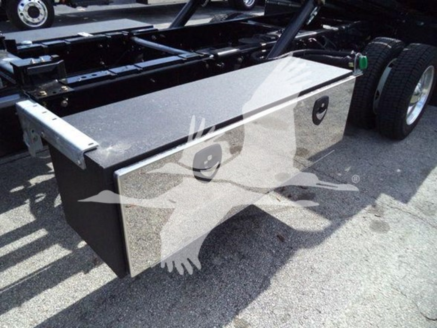 Truck chassis with rectangular metal toolbox mounted under the frame near the rear wheels, view of side showing toolbox lid and latch.