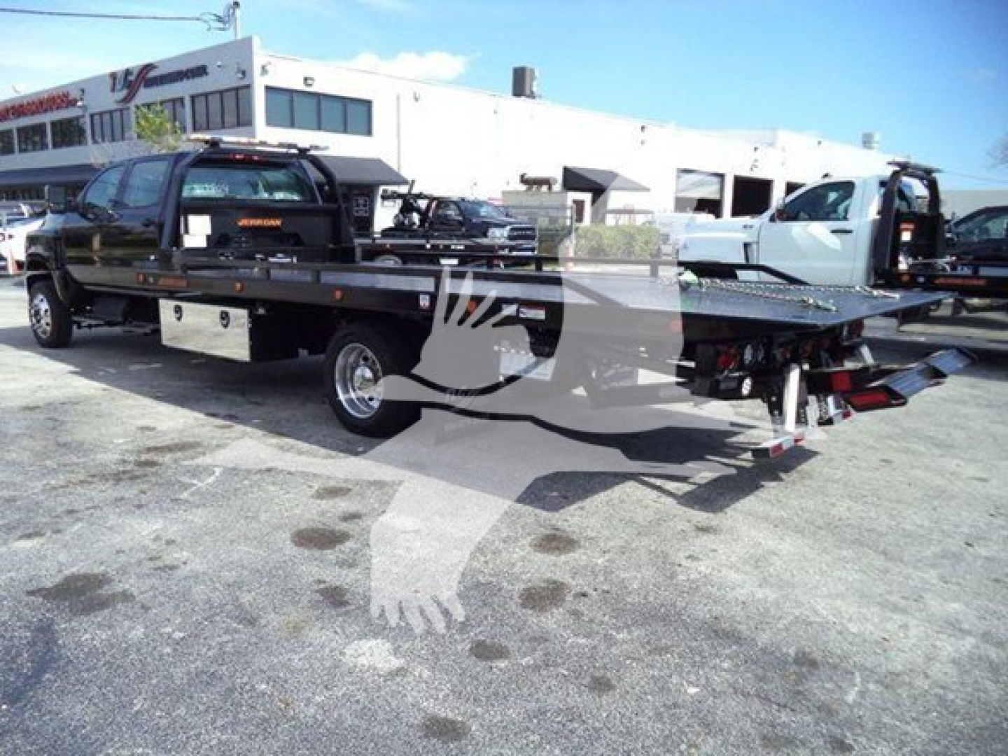 Flatbed tow truck with a black company decal on the cab and a chrome toolbox; the truck has a long flatbed with a hydraulic tilt and a white building in the background.