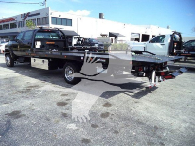 Flatbed tow truck with a black company decal on the cab and a chrome toolbox; the truck has a long flatbed with a hydraulic tilt and a white building in the background.