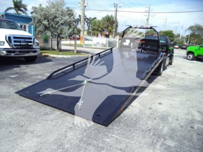 Truck ramp/flatbed on a rollback bed with a 2023 International CV title, showing a lowered steel deck and winch. Side cab visible with other trucks in lot.