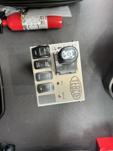 Dashboard control panel with multiple black rocker switches and a round dial, label says FCFBD? and a small red emergency release lever nearby.