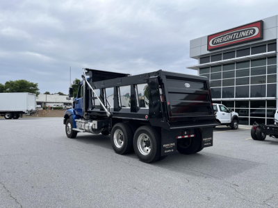 Truck bed of a new Western Star 47X dump truck seen from rear three-quarter view, black dump body, dual rear axles, Freightliner dealership in background.