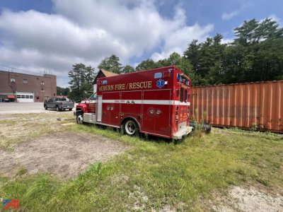 A red Auburn Fire Rescue GMC C7500 fire truck with equipment compartments, emergency lights, and 'Auburn Fire / Rescue' lettering parked outdoors; side view showing wheel and ladder bay.