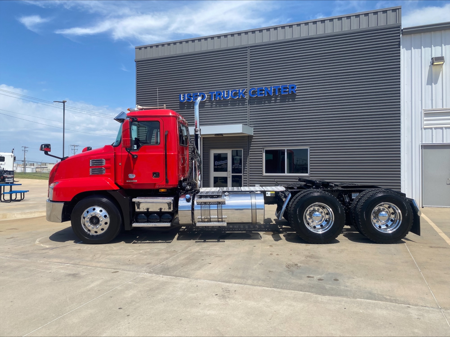 Truck side view of a red 2021 Mack Anthem conventional daycab parked in front of a building labeled US TRUCK CENTER, featuring chrome fuel tanks and a three-axle rear configuration.