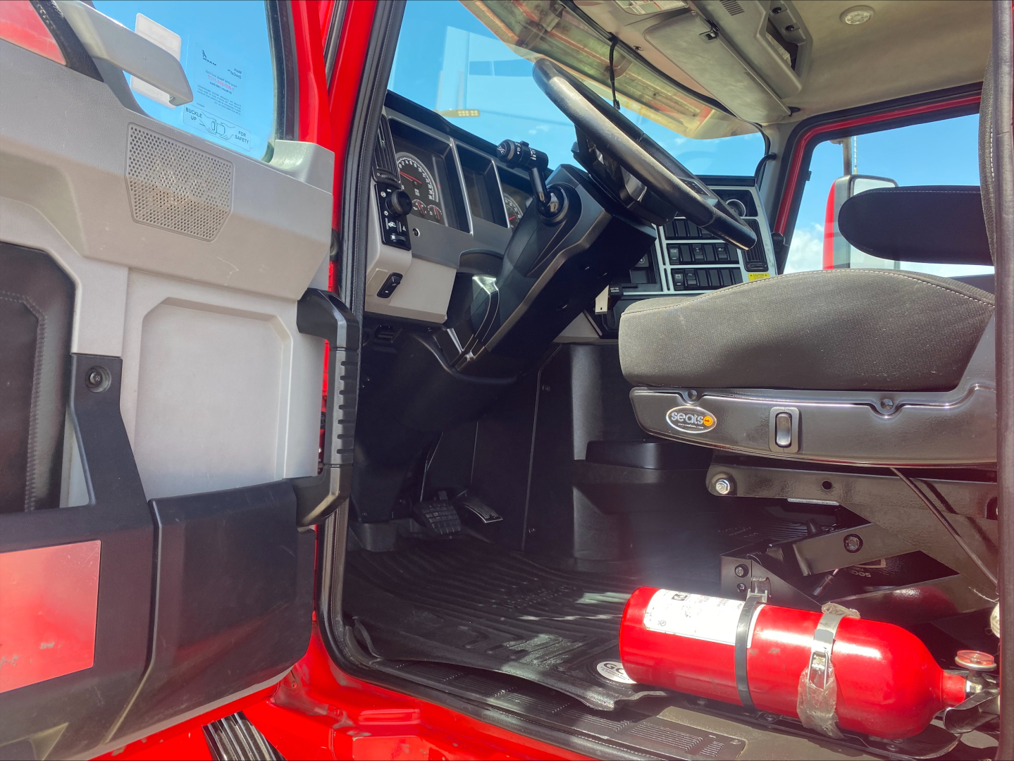 Cab interior of a 2021 Mack Anthem daycab showing steering wheel, driver seat, dashboard, and a mounted fire extinguisher under the dash.