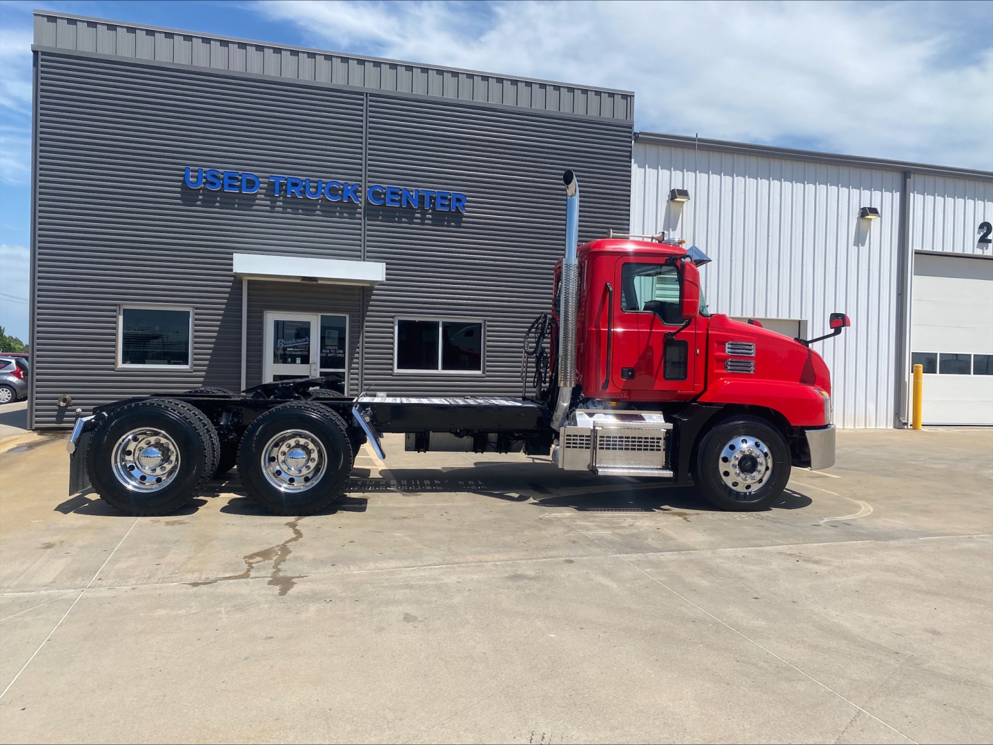 Used Mack truck with red cab and long chassis in front of Used Truck Center building. Visible text includes 'USED TRUCK CENTER' on the building, and no other legible labels on truck.